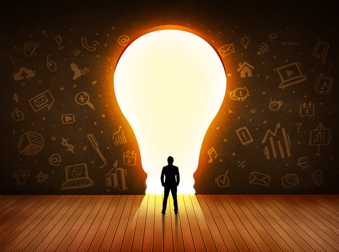 Silhouette of a business professional looking at an entrance shaped like a bright light bulb through a brown wall. The person stands on a laminated wooded floor, and the wall has many business-inspired icon drawings.