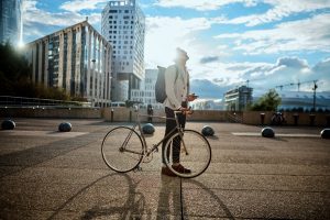 A student is looking into the bright blue sunny sky with a backpack on his shoulders, holding a mobile in one hand and resting the other on a bike. The student is in the city with high glassed buildings. Business Impact article for Leading change and inspiring lifelong learning.