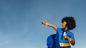 A kid with short black afro hair punching into the sky. The young kid is in a blue and gold superhero outfit with a mask, cap and a lightning bolt icon on his t-shirt. This is to symbolise transformation and impact.