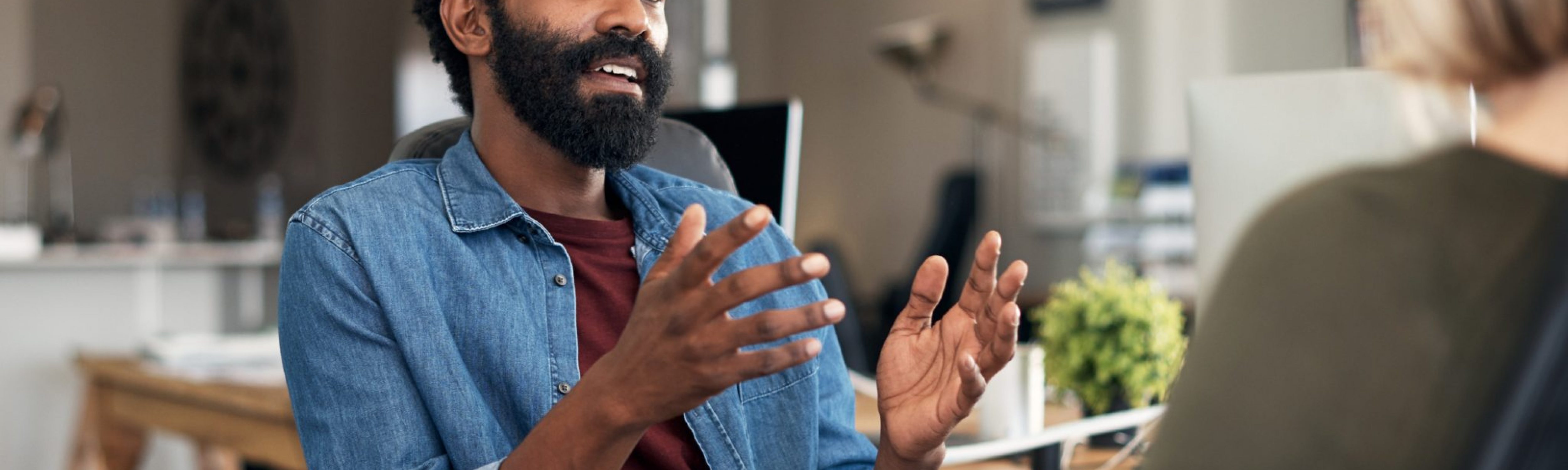 Two casually dressed business individuals in a closed meeting, discussing feedback and planning in a creative office environment. The individual has short black afro hair with a black beard dressed in a maroon tee shirt and a denim long-sleeved on top.