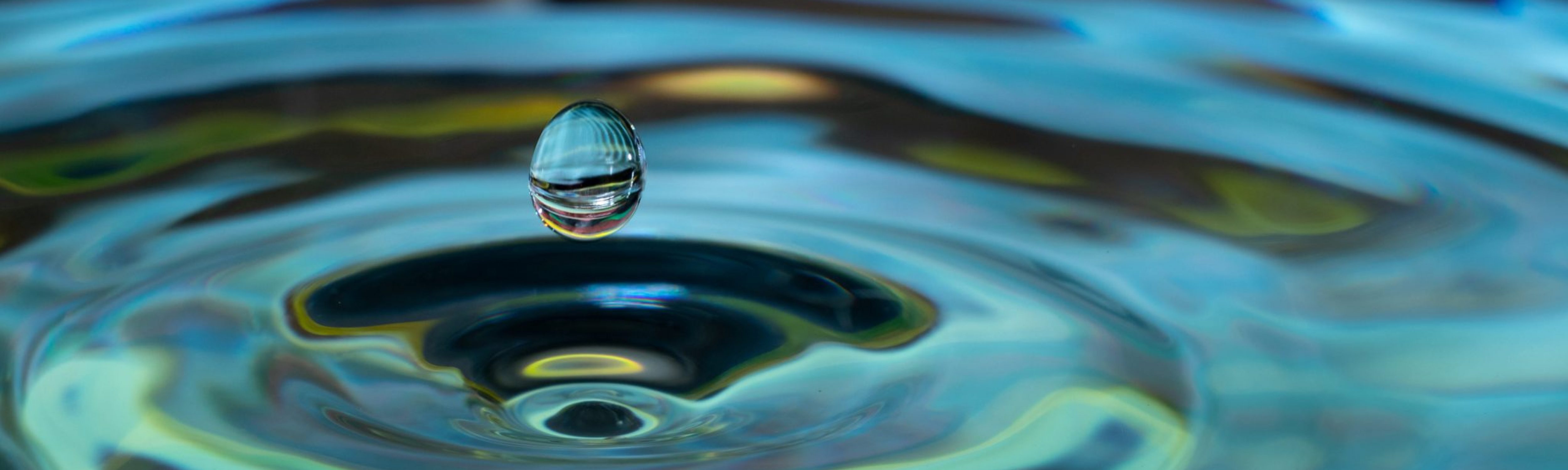 A close-up photograph of a single water droplet about to hit a pool of water; pool of water is causing a circular ripple motion. Water, in this case, signifies sustainability. Business Impact article for Making an impact: corporate social responsibility.